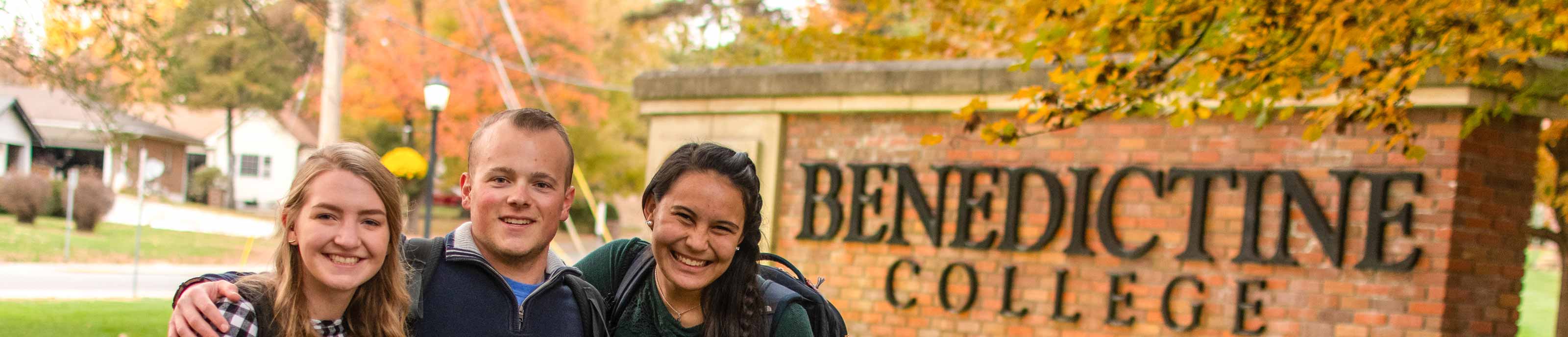 Three students pose for a photo in front of the Benedictine College entrance sign