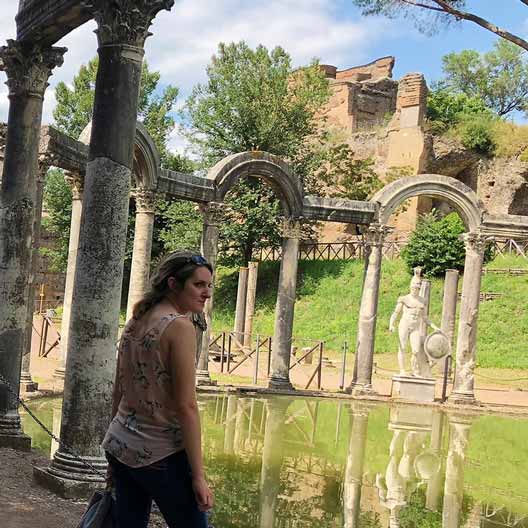 A student stands at a historical site, with a set of arches around a pool of water and a statue in the background