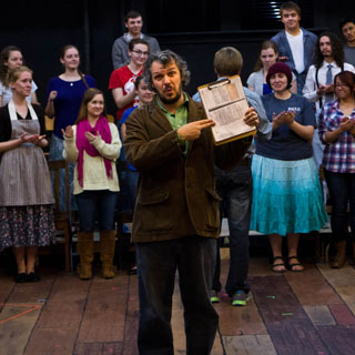 Dr. Scott Cox points at a clipboard on a theatre set with actors in the background