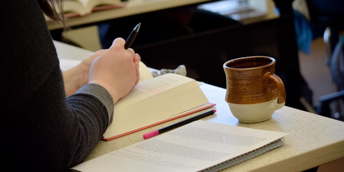 Student sitting at a desk with a book, notes, and coffee mug